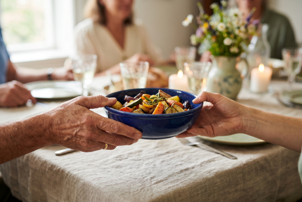 Family dinner traditions — two hands passing a blue ceramic bowl of roasted vegetables across a softly lit table. 