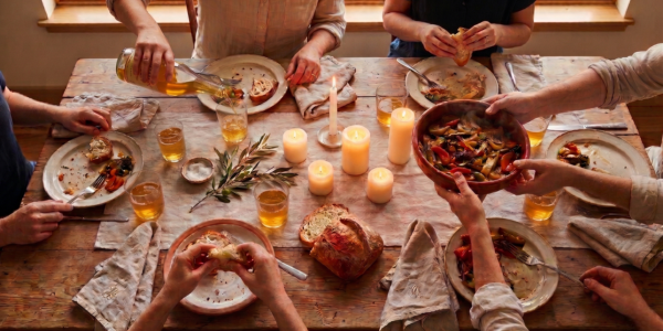Cultural meaning of eating together — hands sharing bread and wine across a rustic wooden dinner table at sunset.