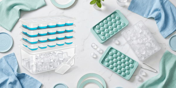Flat lay of silicone ice cube trays and sphere ice molds with a clear storage bin, scoop, and tongs on a marble background.